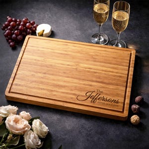 Peut inclure: Planche à découper rectangulaire en bois avec une rainure à jus, gravée de "The Jeffersons ESTABLISHED 2026" en écriture cursive. La planche est entourée de raisins, de fromage, de fleurs et de coupes de champagne.