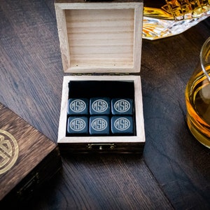 Pu&ograve; includere: Un set di sei pietre per whisky in steatite nera in una scatola di legno con un monogramma dorato. Le pietre sono incise con le iniziali "MSB". La scatola &egrave; aperta e le pietre sono visibili all'interno.