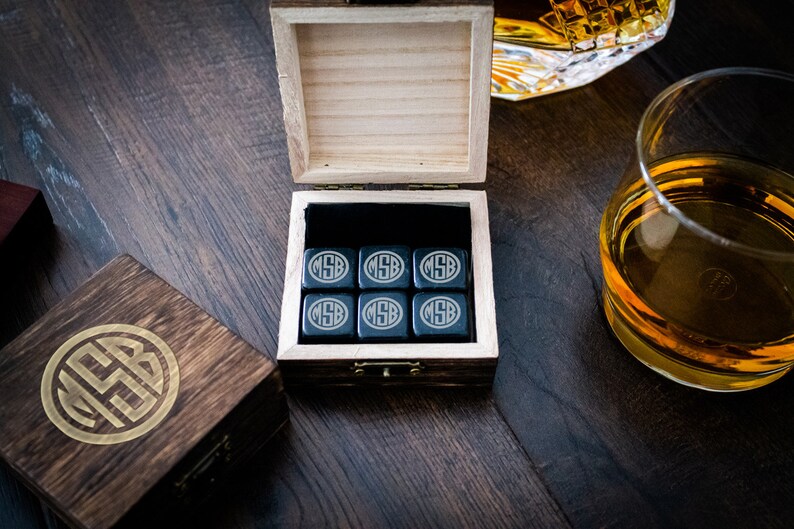 Pu&ograve; includere: Un set di sei pietre per whisky in steatite nera in una scatola di legno con un monogramma dorato. Le pietre sono incise con le iniziali "MSB". La scatola &egrave; aperta e le pietre sono visibili all'interno.