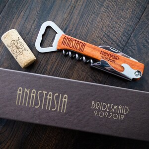 May include: A wooden bottle opener and corkscrew with the name "ANASTASIA" engraved on the handle. The words "Bridesmaid 9.09.2019" are also engraved on the handle. A cork and a brown box with the name "ANASTASIA" and "Bridesmaid 9.09.2019" are also visible.