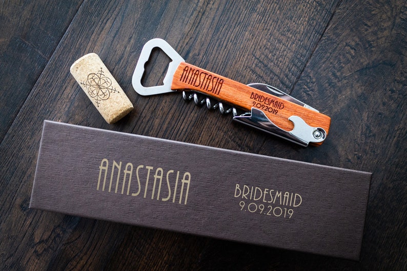 May include: A wooden bottle opener and corkscrew with the name "ANASTASIA" engraved on the handle. The words "Bridesmaid 9.09.2019" are also engraved on the handle. A cork and a brown box with the name "ANASTASIA" and "Bridesmaid 9.09.2019" are also visible.