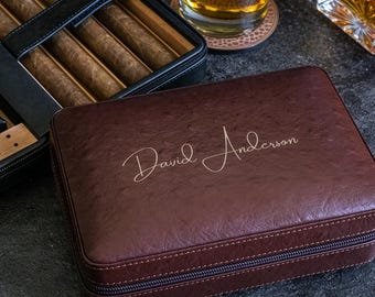 May include: A brown leather cigar case with the name "David Anderson" in a cursive font. The case is closed with a zipper and sits next to an open black cigar case filled with cigars. A glass of amber liquid is in the background.