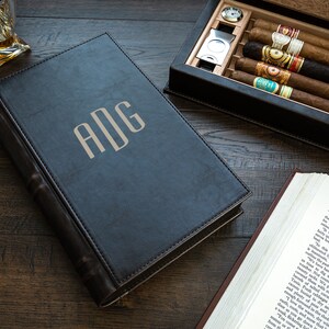 May include: A black leather-bound cigar box with a monogram "ADG" on the front. The box is open and contains several cigars. The box is sitting on a wooden table.
