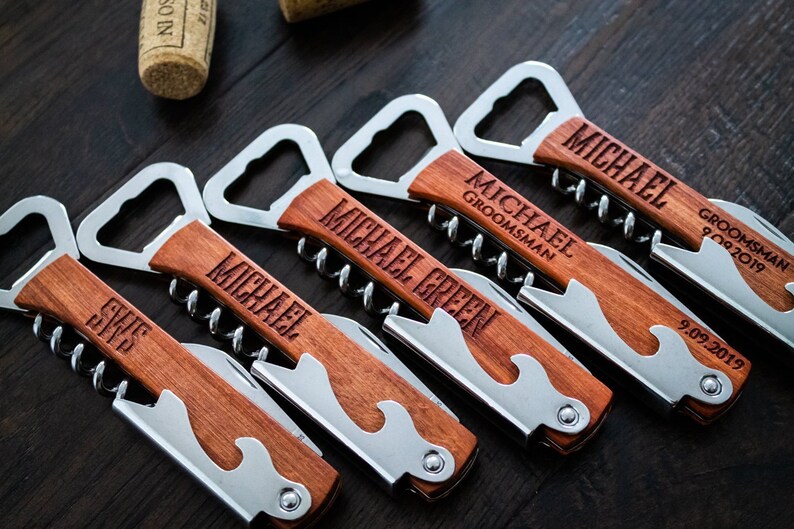May include: A collection of personalized wooden bottle openers and corkscrews. Each opener has a stainless steel bottle opener, corkscrew, and knife. The wooden handles are engraved with names and dates, perfect for a wedding or special event.