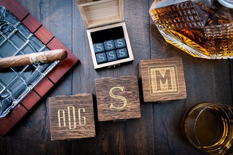 Pu&ograve; includere: Tre scatole di legno con iniziali incise, "ADG", "S" e "M". &Egrave; anche mostrata una scatola con sei cubetti di pietra nera incisi con la lettera "S". Sono anche mostrati una decanter di vetro con liquido ambrato e un bicchiere con liquido ambrato.