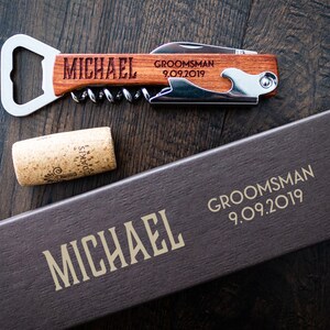 May include: A wooden bottle opener and corkscrew with the name "MICHAEL" engraved on the handle. The words "GROOMSMAN 9.09.2019" are also engraved. A cork and a box with the name "MICHAEL" and "GROOMSMAN 9.09.2019" are also visible.