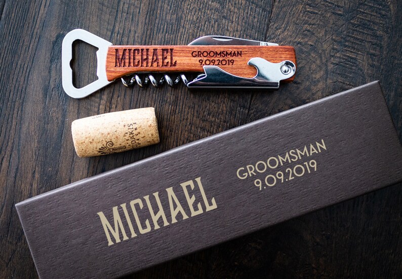 May include: A wooden bottle opener and corkscrew with the name "MICHAEL" engraved on the handle. The words "GROOMSMAN 9.09.2019" are also engraved. A cork and a box with the name "MICHAEL" and "GROOMSMAN 9.09.2019" are also visible.