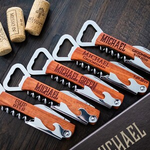 May include: Wooden corkscrews with stainless steel accents, personalized with names and dates. The corkscrews have a bottle opener, knife, and corkscrew. The image also shows wine corks and a box with text.