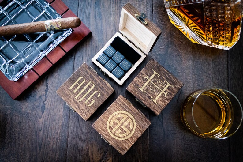 Pu&ograve; includere: Quattro scatole di legno con iniziali incise e un set di pietre per whisky nere in una scatola di legno con fodera in velluto nero. Le scatole sono su una superficie in legno scuro con un bicchiere di whisky e un posacenere per sigari.