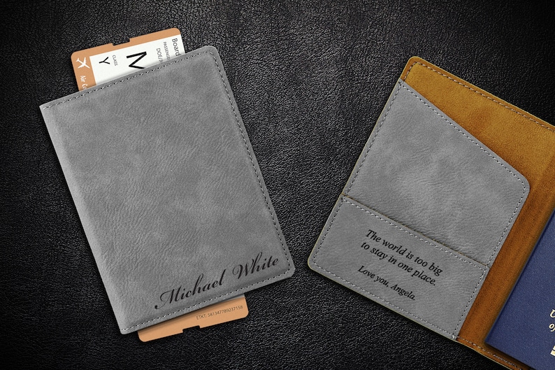 Puede incluir: Dos porta pasaportes grises con detalles marrones. Uno tiene el nombre "Michael White" grabado. El otro tiene el texto "El mundo es demasiado grande para quedarse en un solo lugar. Te quiero, Angela." grabado.