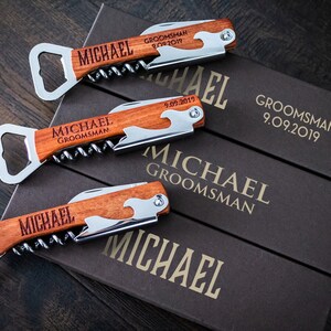 May include: Three personalized wooden corkscrews with bottle opener and knife features, engraved with "MICHAEL GROOMSMAN 9.09.2019". The corkscrews are displayed on top of three brown boxes with the same text.
