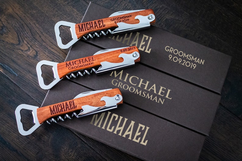 May include: Three personalized wooden corkscrews with bottle opener and knife features, engraved with "MICHAEL GROOMSMAN 9.09.2019". The corkscrews are displayed on top of three brown boxes with the same text.
