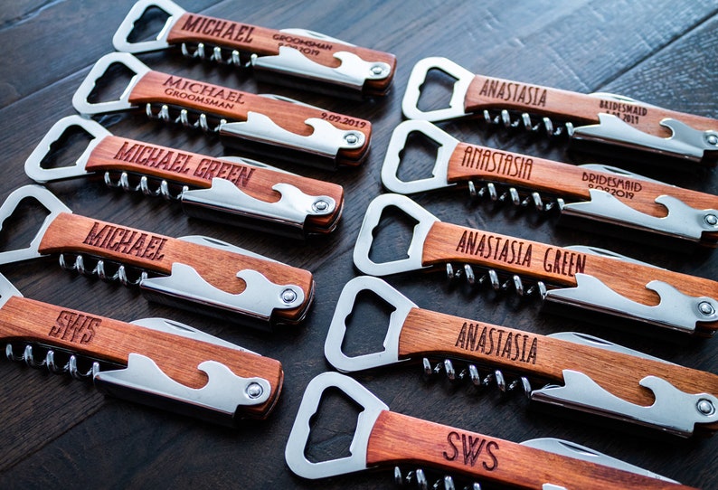 May include: A collection of personalized wooden bottle openers with corkscrews and bottle openers. Each opener has a stainless steel blade and is engraved with names and titles like "Groomsman" and "Bridesmaid".