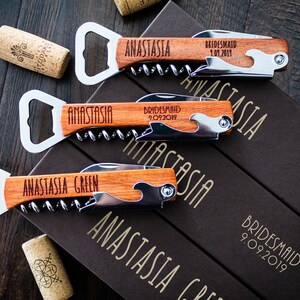 May include: Wooden bottle openers with corkscrews and bottle openers, engraved with names and dates. The openers are lying on a dark wooden surface, with corks and boxes visible. The text "ANASTASIA" and "BRIDESMAID" are visible.