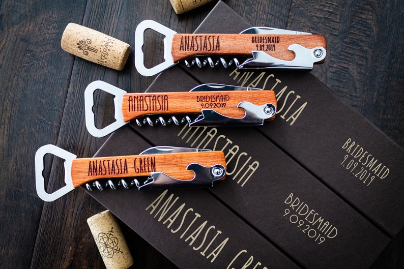 May include: Wooden bottle openers with corkscrews and bottle openers, engraved with names and dates. The openers are lying on a dark wooden surface, with corks and boxes visible. The text "ANASTASIA" and "BRIDESMAID" are visible.