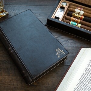 May include: A black leather-bound book with a gold embossed letter "A" on the cover. The book is closed and lying on a wooden surface. There is a cigar box with cigars and a glass of whiskey in the background.