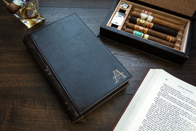 May include: A black leather-bound book with a gold embossed letter "A" on the cover. The book is closed and lying on a wooden surface. There is a cigar box with cigars and a glass of whiskey in the background.