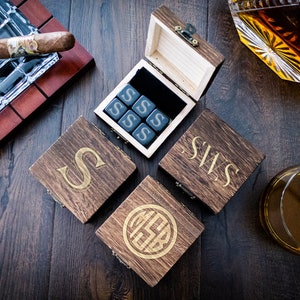 Pu&ograve; includere: Un set di quattro scatole di legno con iniziali incise. Ogni scatola contiene un set di dadi di pietra nera con la lettera "S" incisa su ogni lato. Le scatole sono disposte su una superficie di legno scuro con un bicchiere di whisky, un sigaro e un posacenere.