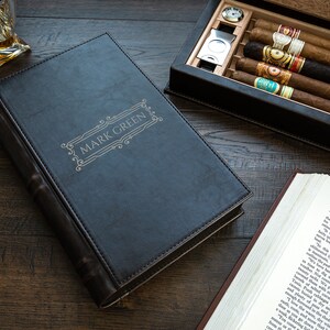 May include: A black leather-bound cigar box with the name "MARK GREEN" embossed on the front. The box is open and contains several cigars and a cigar cutter.
