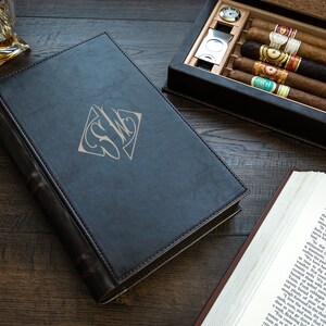 May include: A black leather-bound book with a gold monogram "S.W.C." on the cover. The book is closed and lying on a wooden surface. There is a cigar box with cigars and a glass of whiskey in the background.