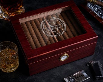 May include: A mahogany cigar humidor with a glass top, filled with cigars. The humidor has a personalized engraving that reads "Sanders" inside a laurel wreath. A cigar cutter, lighter, and a glass of whiskey are also visible.