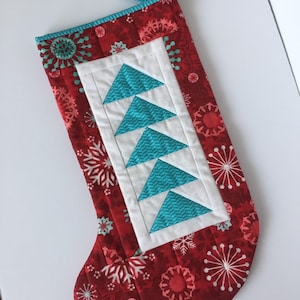 May include: A red Christmas stocking with a white and turquoise quilted design of a Christmas tree. The stocking has a white cuff and a turquoise and white loop at the top.