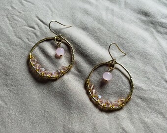 May include: A pair of gold-toned hoop earrings. Each earring features a small, light pink bead suspended from the top and a row of pink crystal beads along the bottom half of the hoop. The earrings are set against a neutral background.