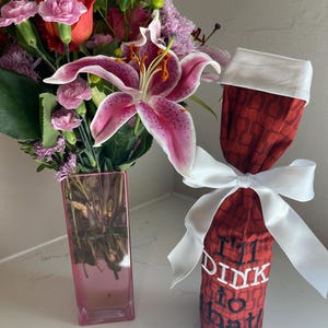 May include: A bouquet of flowers in a pink rectangular vase next to a red wine bottle bag. The bouquet includes pink lilies, roses, and other flowers. The wine bag has the text "I'll Dink to that" and is tied with a white ribbon.