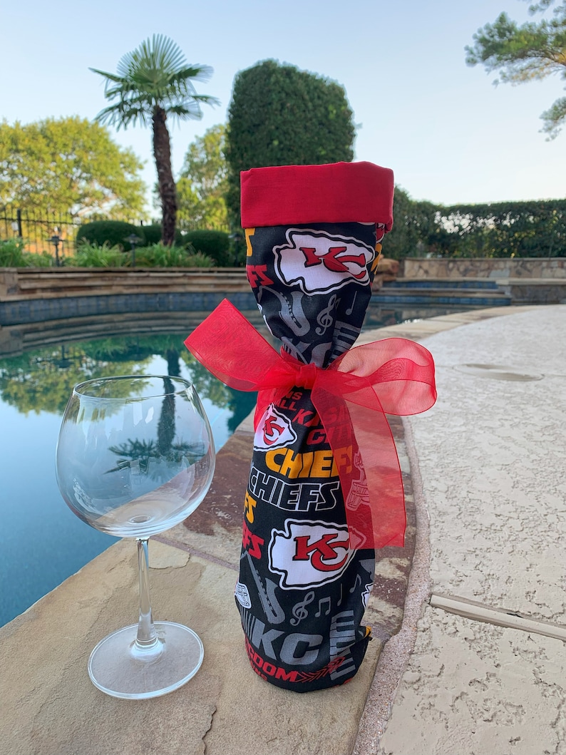 May include: A black and red wine bottle bag with the Kansas City Chiefs logo and the words "Chiefs" and "Kingdom". The bag is tied with a red ribbon and is sitting on a concrete surface next to a clear wine glass.
