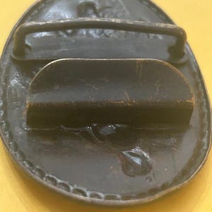 May include: A vintage oval-shaped belt buckle with a dark bronze finish. The buckle features a raised design around the edge and a bar for the belt loop. The back has a curved bar and a pin for securing the belt.