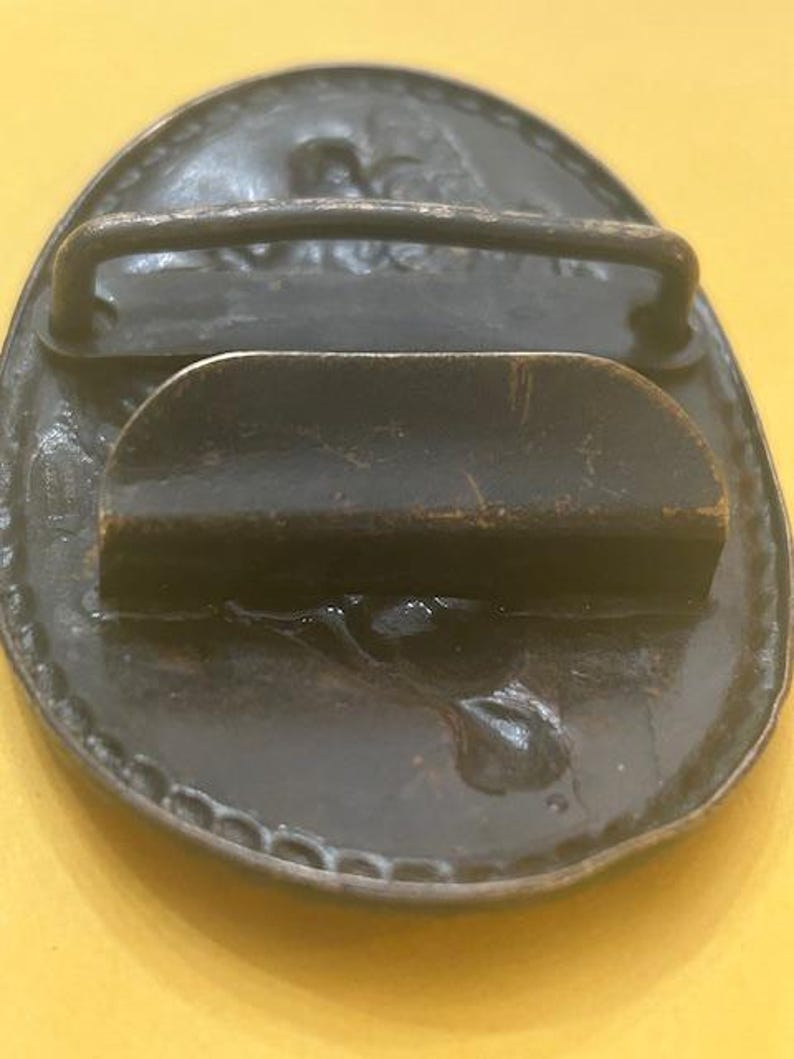 May include: A vintage oval-shaped belt buckle with a dark bronze finish. The buckle features a raised design around the edge and a bar for the belt loop. The back has a curved bar and a pin for securing the belt.