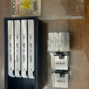 May include: A collection of RIDGID brand plumbing tools. The image shows a black case containing four metal inserts labeled 1-4, and several other metal pieces. A clear plastic case with a label that reads "RIDGID GENUINE PARTS" is also visible.