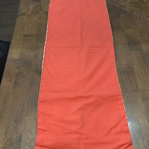 Puede incluir: Un camino de mesa rectangular en color naranja coral s&oacute;lido. El camino presenta una superficie de tela lisa y plana con bordes rectos y acabados a lo largo de los lados y extremos, ofreciendo un dise&ntilde;o limpio y sencillo para la decoraci&oacute;n.