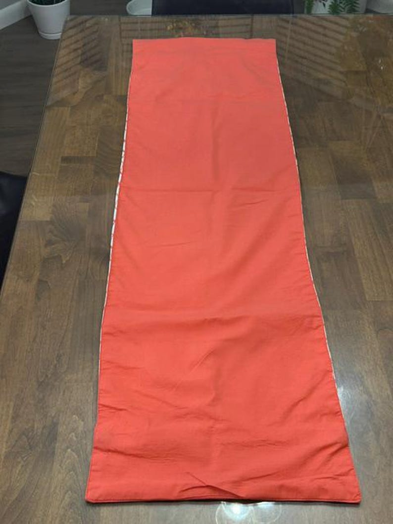 Puede incluir: Un camino de mesa rectangular en color naranja coral s&oacute;lido. El camino presenta una superficie de tela lisa y plana con bordes rectos y acabados a lo largo de los lados y extremos, ofreciendo un dise&ntilde;o limpio y sencillo para la decoraci&oacute;n.