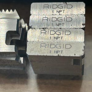 May include: Close-up of several metal RIDGID 1 NPT die heads. The die heads are stacked and have a silver finish. The text "RIDGID" and "1 NPT" are engraved on the top of each die head. The die heads are used for threading pipes.