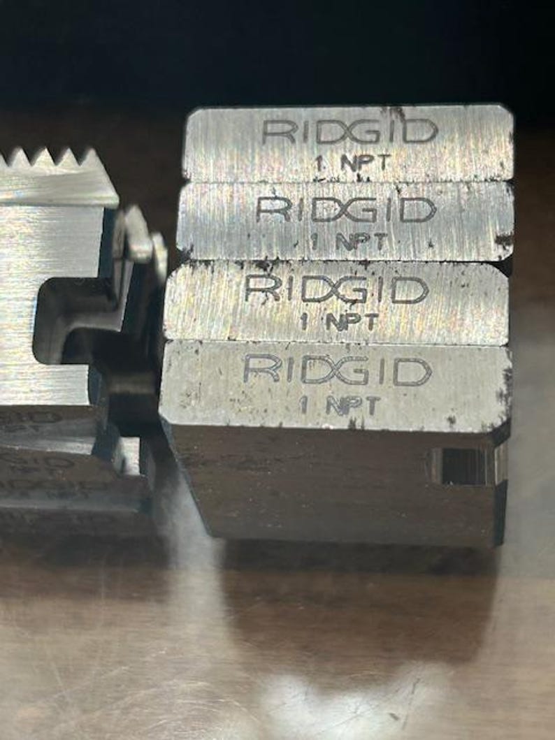May include: Close-up of several metal RIDGID 1 NPT die heads. The die heads are stacked and have a silver finish. The text "RIDGID" and "1 NPT" are engraved on the top of each die head. The die heads are used for threading pipes.