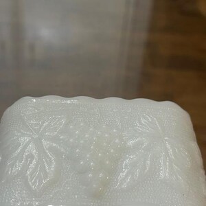 May include: A rectangular white milk glass dish featuring a raised, embossed pattern of grape clusters and vine leaves. The edges of the dish have a scalloped design, and the background texture of the glass is finely stippled throughout.