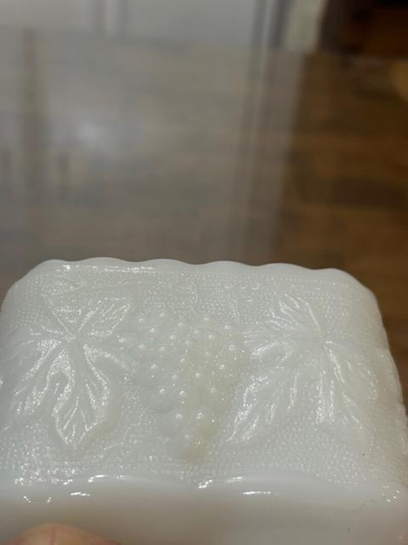 May include: A rectangular white milk glass dish featuring a raised, embossed pattern of grape clusters and vine leaves. The edges of the dish have a scalloped design, and the background texture of the glass is finely stippled throughout.