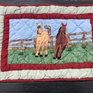 May include: A quilted throw pillow with a green, brown, and blue scene of two horses in a field with a wooden fence. The pillow has a red and white border.