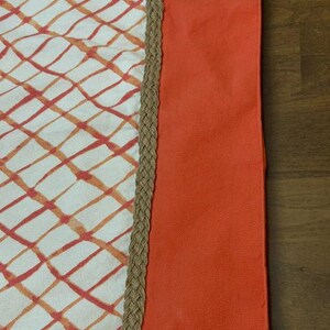 Puede incluir: Un camino de mesa rectangular con base blanca y patr&oacute;n de cuadr&iacute;cula naranja. Un borde grueso de yute trenzado en color tostado separa la secci&oacute;n estampada de un borde s&oacute;lido de color naranja vibrante a lo largo del lado derecho.