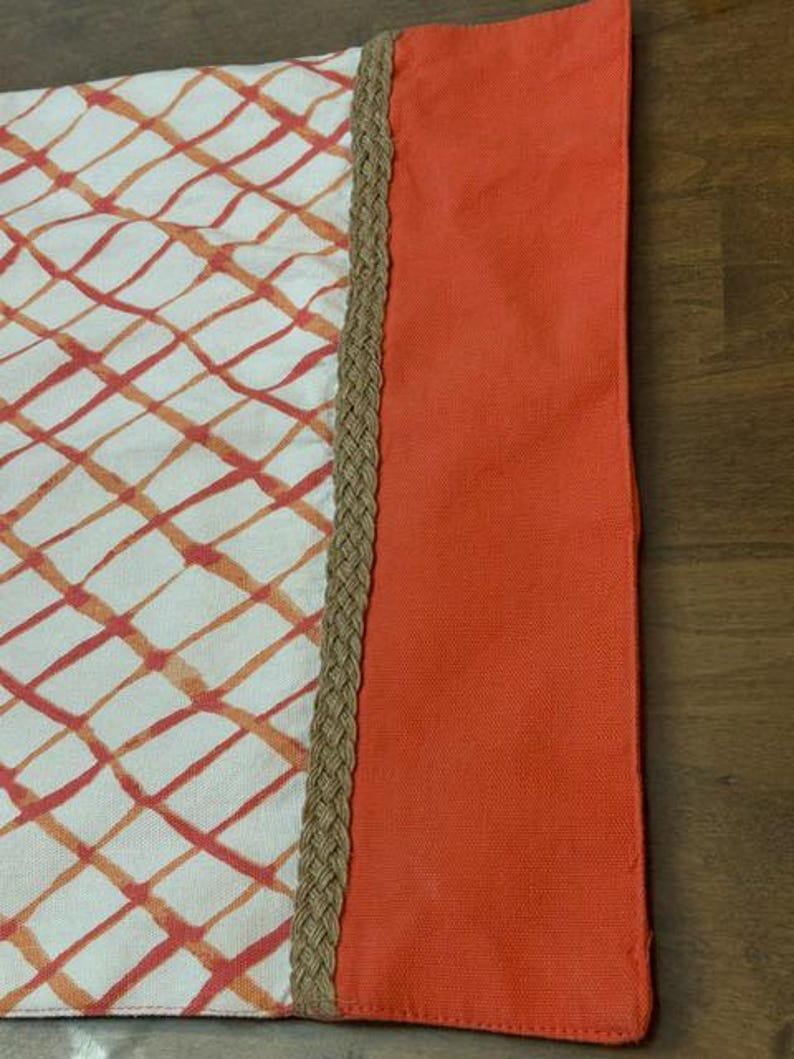 Puede incluir: Un camino de mesa rectangular con base blanca y patr&oacute;n de cuadr&iacute;cula naranja. Un borde grueso de yute trenzado en color tostado separa la secci&oacute;n estampada de un borde s&oacute;lido de color naranja vibrante a lo largo del lado derecho.