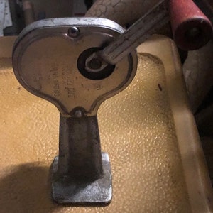 May include: A vintage metal hand crank with a red handle. The crank has a metal base and a silver handle. The crank is labeled "THE LITTLE WONDER 0054300".