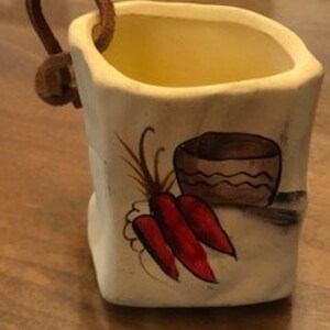 May include: A small, white ceramic planter with a rustic, textured finish. It features a hand-painted design of red carrots and a brown pot with wavy lines. A brown leather strap with a wooden toggle serves as a handle.