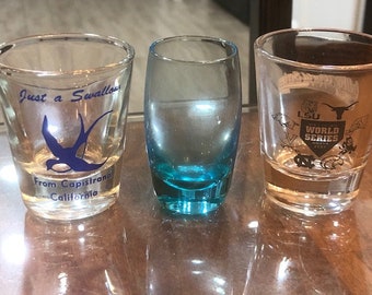 May include: Three clear glass shot glasses. The left glass has a blue swallow design and the text "Just a Swallow from Capistrano California". The middle glass has a blue liquid and a light blue bottom. The right glass has a black and white design with the text "LSU World Series 2009".