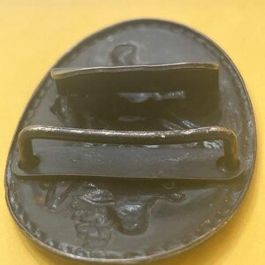 May include: A dark metal belt buckle with an oval shape. The buckle has a raised design and a bar for the belt to go through. The buckle is set against a yellow background.