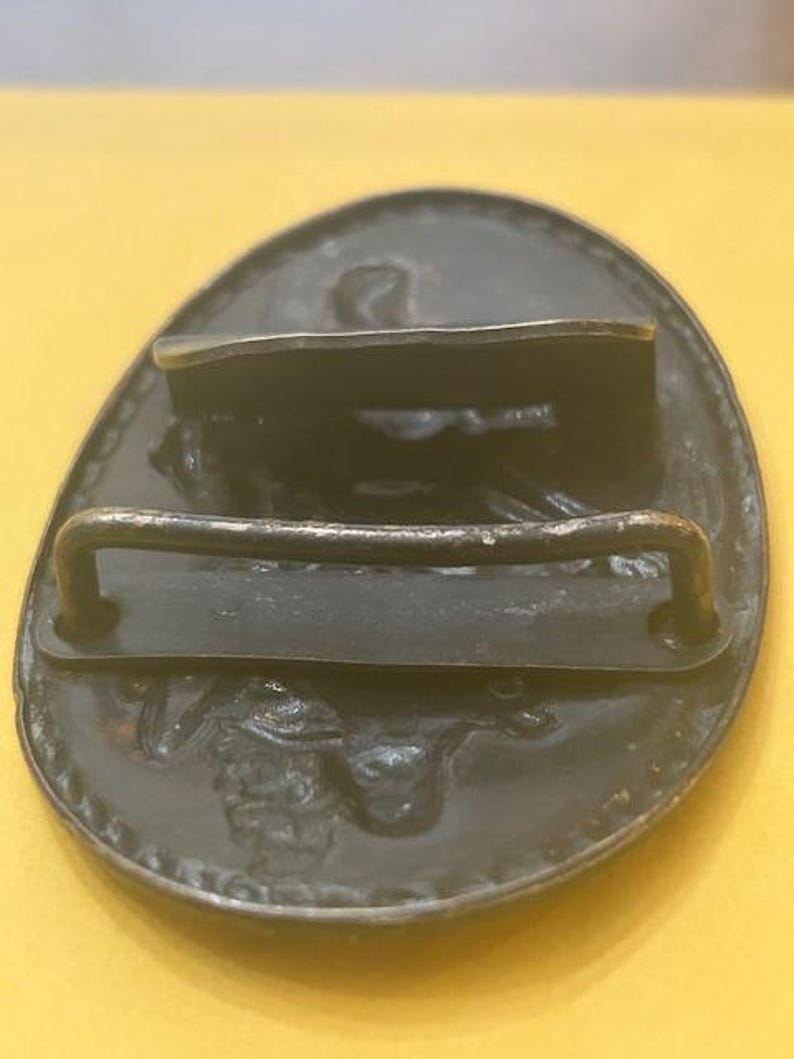 May include: A dark metal belt buckle with an oval shape. The buckle has a raised design and a bar for the belt to go through. The buckle is set against a yellow background.