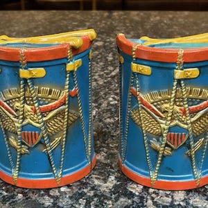 May include: Two blue, red, and yellow drum-shaped salt and pepper shakers. Each shaker features a gold eagle emblem and drumsticks. The shakers are cylindrical with a red rim and base.