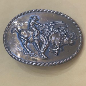 May include: Oval-shaped silver-toned belt buckle featuring a cowboy riding a bucking bronco, roping a bull. The buckle has a rope-like border and is likely made of metal. A classic Western-themed accessory.