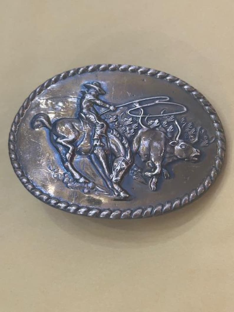 May include: Oval-shaped silver-toned belt buckle featuring a cowboy riding a bucking bronco, roping a bull. The buckle has a rope-like border and is likely made of metal. A classic Western-themed accessory.