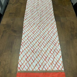 Puede incluir: Un camino de mesa rectangular con un panel central blanco que presenta un patr&oacute;n de cuadr&iacute;cula cruzada en naranja y rojo. El camino est&aacute; bordeado en ambos extremos por un ribete trenzado de color tostado y una banda de tela naranja s&oacute;lida.